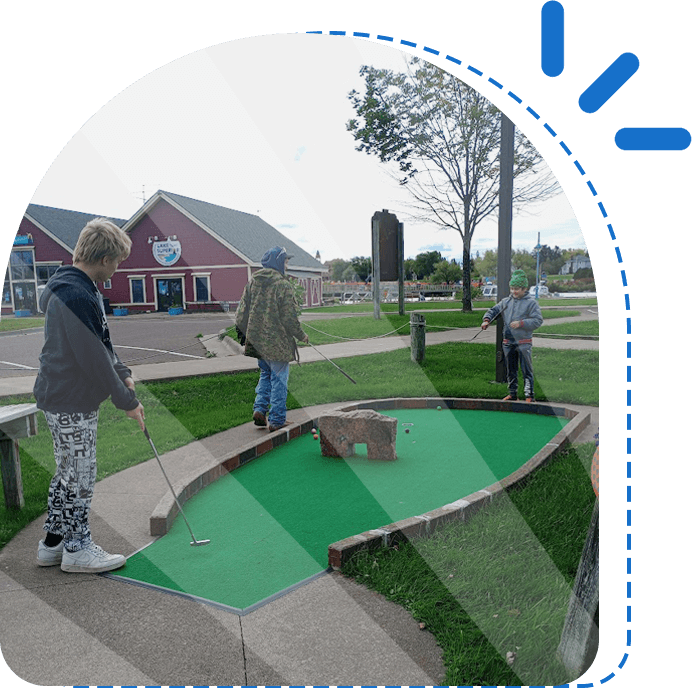 Three people, each with a golf putter, playing mini golf on a green course with obstacles, in a suburban setting.