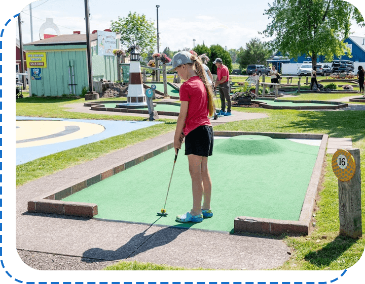A woman playing mini-golf at an outdoor course with other players in the background on a sunny day.